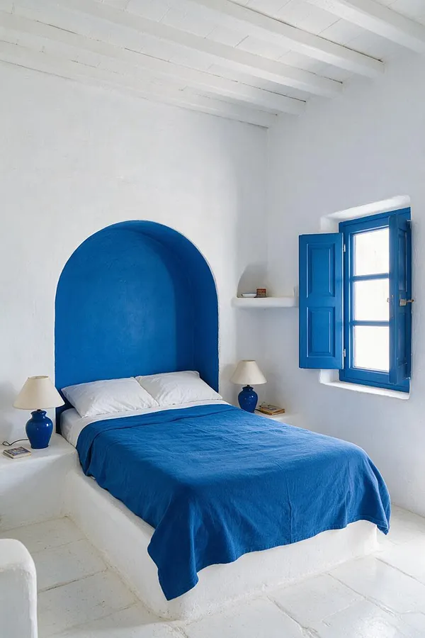 A bright Mediterranean-style bedroom with whitewashed walls, a curved cobalt blue headboard niche, and a matching blue bedspread. Blue shuttered windows, simple white bedding, and small bedside lamps create a calm, sunlit, minimalist space.