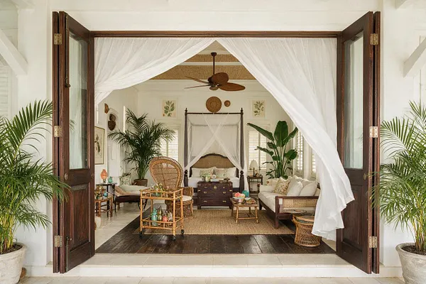An open set of dark wood French doors reveals a bright tropical living room framed by flowing white sheer curtains. Inside are rattan and wicker furniture, a canopy bed at the back, large potted palms, white walls, woven textures, and warm wood floors in a relaxed, airy setting.