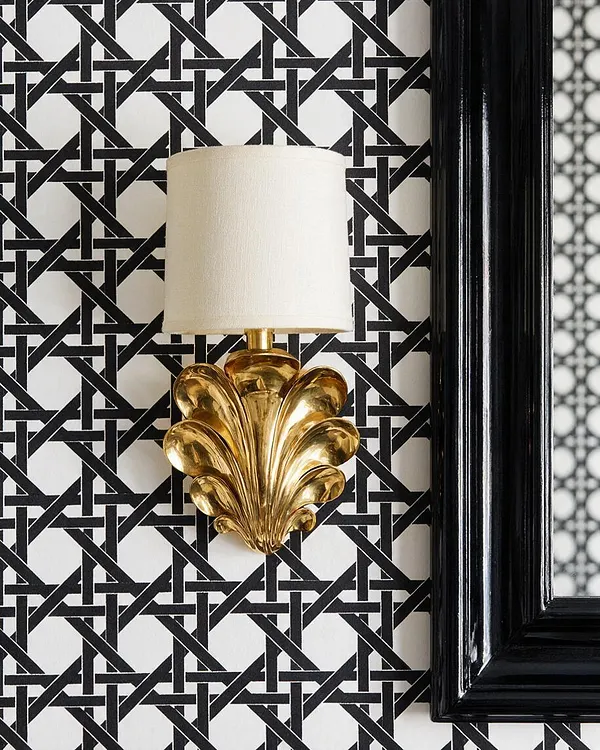 A close-up of a Hollywood Regency wall sconce with a white cylindrical shade and a polished gold shell-shaped base mounted on black-and-white geometric wallpaper. A glossy black framed mirror or trim appears along the right edge, adding a dramatic contrast.