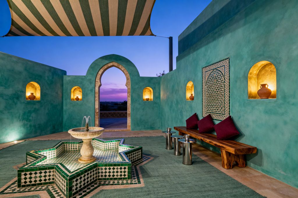 An open-air Moroccan patio at dusk with turquoise tadelakt walls and a limestone horseshoe arch, centered by an eight-pointed star fountain clad in green-and-white zellige and topped by a marble bowl. Gold-toned wall niches holding terracotta pots, a live-edge wooden bench with burgundy cushions, polished metal side tables, a sage flatweave rug, and a striped canvas canopy create a warm, intimate setting.