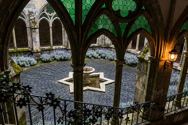 View from under pointed stone arches with green circular-paned stained glass, overlooking a cloistered patio. At the center, a carved stone fountain sits within an eight-pointed star basin, set in radial charcoal slate paving; borders of lavender and silvery shrubs ring the court as weathered limestone columns and tracery frame the scene, while a wrought-iron lantern casts warm light against moss-covered walls.