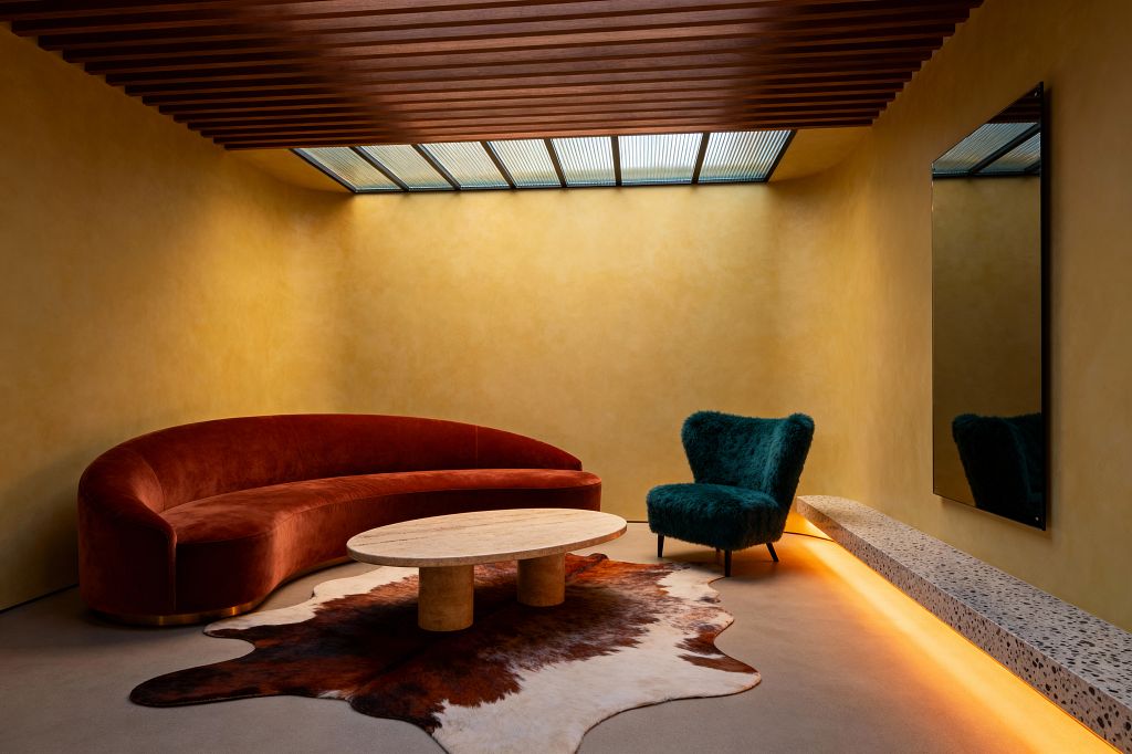 A contemporary living room with ochre plaster walls and a ribbed wood-slat ceiling features a linear skylight above a curved rust velvet sofa, an oval travertine coffee table with cylindrical legs, and a brown-and-white cowhide rug. A deep teal, fuzzy lounge chair sits beside a floating terrazzo bench with warm amber underlighting, while a large black-framed mirror and smooth concrete floor complete the minimalist, sculptural setting.