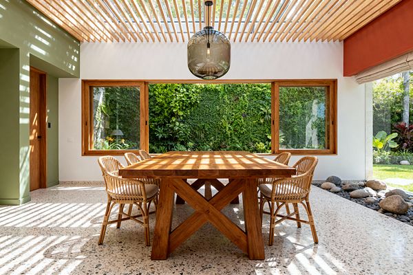 A sunlit dining room features a thick solid-wood table and six rattan chairs on speckled terrazzo flooring. A skylight with timber slats casts striped shadows, a smoked-glass pendant hangs centrally, large wood-framed windows face a lush living wall and garden, and a river-stone border lines the edge beside white plaster, a sage-painted wall, and a terracotta soffit.