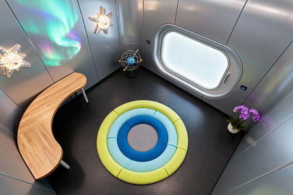A compact kids’ room styled like a spaceship features brushed metal wall panels, a rounded rectangular frosted porthole window, and black coin-textured flooring. At center sits a circular layered rug in lime, aqua, cobalt, and gray; a crescent oak desk hugs the wall, star-shaped wall sconces and an armillary globe lamp provide playful lighting, and a white pot with purple orchids adds a soft natural touch beside aurora-like reflections.