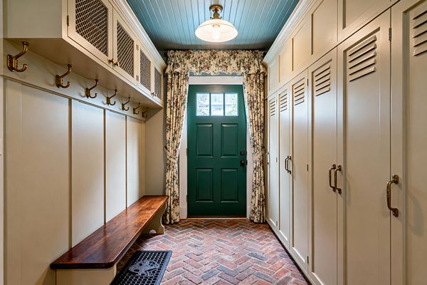 A narrow traditional mudroom with creamy built-in cabinetry: louvered lockers with brass pulls on the right, and a dark-stained wood bench with brass hooks and mesh-front upper cabinets on the left. The red herringbone brick floor leads to a deep green paneled door framed by cream floral curtains, while a muted blue beadboard ceiling and a schoolhouse pendant light create a warm, classic atmosphere.