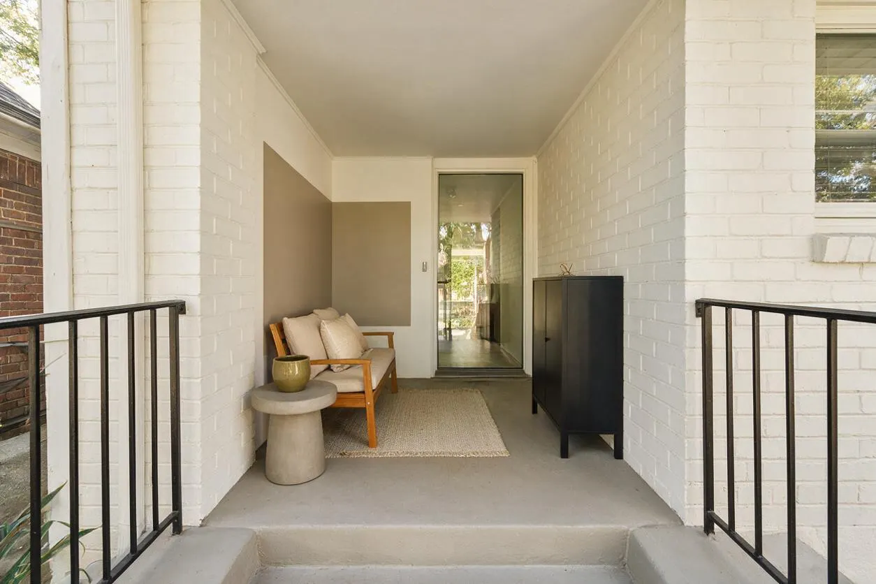 A modern front porch with white painted brick walls, a smooth concrete floor, and black metal railings. A small wooden bench with cream cushions, a round concrete side table, a textured rug, and a slim black storage cabinet create a calm, minimalist seating area.