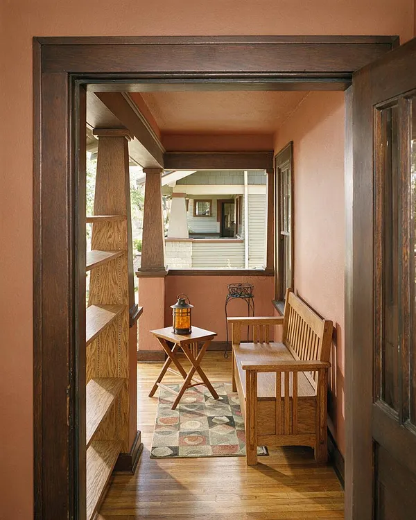 A narrow Craftsman-style front porch with warm peach walls, dark wood trim, built-in shelving, and a slatted wooden bench. A small folding side table with a lantern sits on a patterned rug over wood flooring, creating a cozy, welcoming entryway.