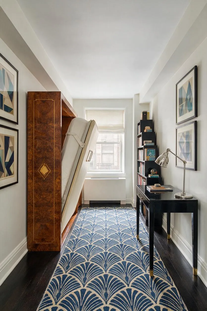 A narrow studio apartment interior with a fold-down bed concealed inside a glossy burl wood cabinet on the left, a black writing desk and shelving on the right, and a blue-and-cream geometric rug running down the center. Framed abstract prints line the walls, while a bright window at the end of the room adds natural light.
