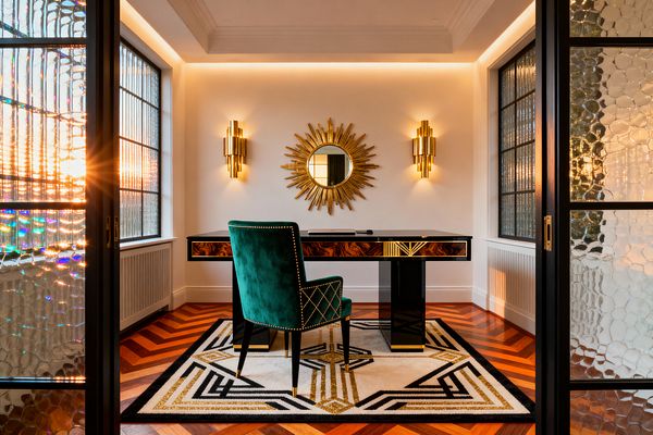 A compact Art Deco home office viewed through textured glass doors features a black lacquer desk with Macassar ebony veneer and brass detailing centered on a cream rug with black and gold geometric motifs. An emerald velvet armchair with brass nailheads faces a white wall hung with a gold sunburst mirror flanked by tiered brass sconces; cove lighting, chevron hardwood flooring, and reeded side windows create a warm, glamorous glow.