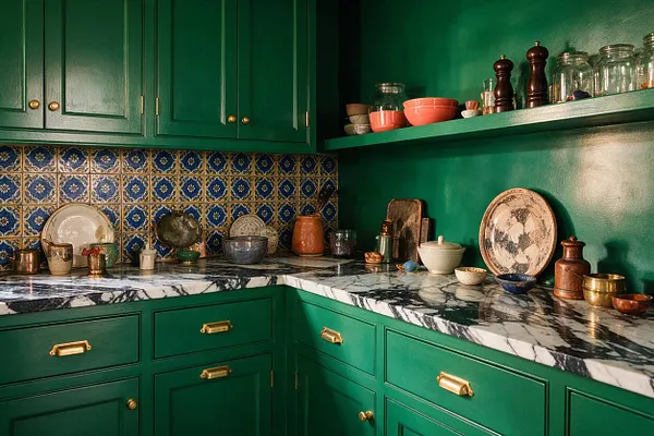 A dramatic kitchen vignette with glossy emerald green cabinets and walls, brass hardware, and a black-and-white marble countertop. The backsplash features ornate blue, gold, and cream patterned tile, while shelves and counters display ceramic bowls, glass jars, copper vessels, and dark wood mills for a layered maximalist look.
