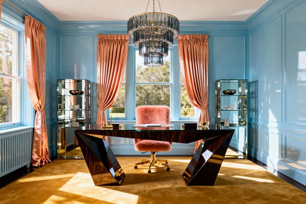 A Hollywood Regency home office with powder-blue lacquered wall paneling features a sculptural, high-gloss dark wood desk centered under a tiered chandelier of smoked glass and brass. Blush-peach satin curtains frame tall windows, mirrored display cabinets flank the desk, a mustard-gold carpet warms the floor, and a pink faux-fur swivel chair on a brass base adds playful glamour.