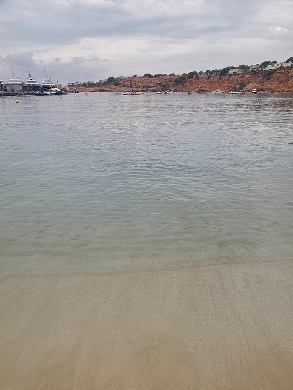 A calm beach scene with clear, shallow water, a sandy shore, yachts docked in a marina, and houses on a cliff under a cloudy sky.