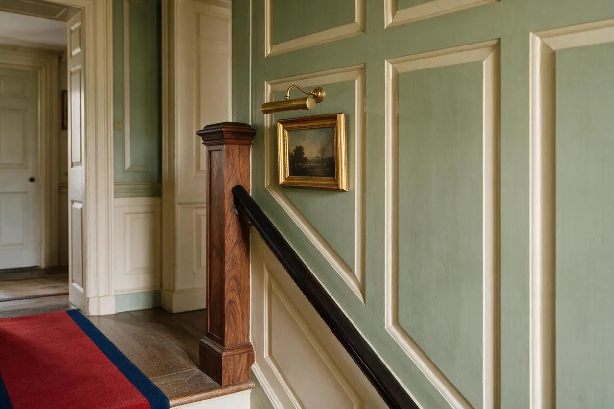 A traditional interior stair hall with pale green paneled walls trimmed in cream, a polished dark wood handrail, and a turned wood newel post. A small framed landscape and brass picture light add classic charm, while a red runner with a blue border anchors the hallway.