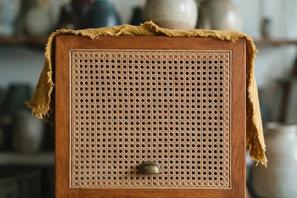 A close-up view of a wooden cabinet front with woven cane paneling and a small round brass knob. A mustard-yellow fringed textile is draped loosely over the top, adding a bohemian layered look against a softly blurred background of pottery and ceramics.
