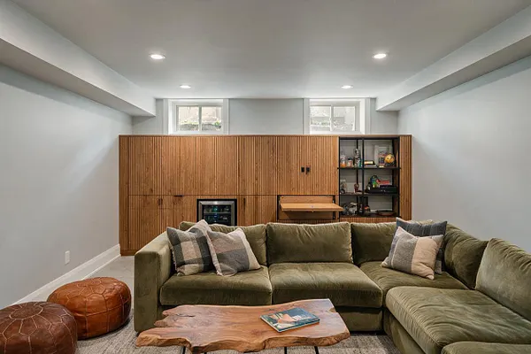A modern basement lounge with a large olive-green sectional sofa, leather poufs, and a natural wood live-edge coffee table. Behind the seating is a full-height slatted wood wall with built-in storage, a small bar area, and a compact beverage fridge beneath recessed ceiling lights.