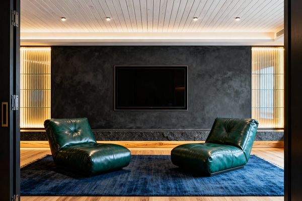 A contemporary media room with two low emerald green leather lounge chairs on a deep blue rug facing a wall‑mounted TV set into a charcoal plaster feature wall. Fluted, backlit glass shelving stands symmetrically on both sides, above a dark stone plinth. The space has light oak flooring and a white plank ceiling with recessed spotlights and warm cove lighting, creating a moody yet inviting atmosphere.