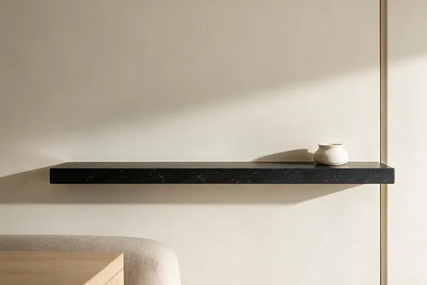 A minimalist interior corner with a slim black stone wall shelf mounted against a warm beige wall. A small white ceramic vase sits on the shelf, while soft natural light casts gentle shadows across the clean, neutral space.
