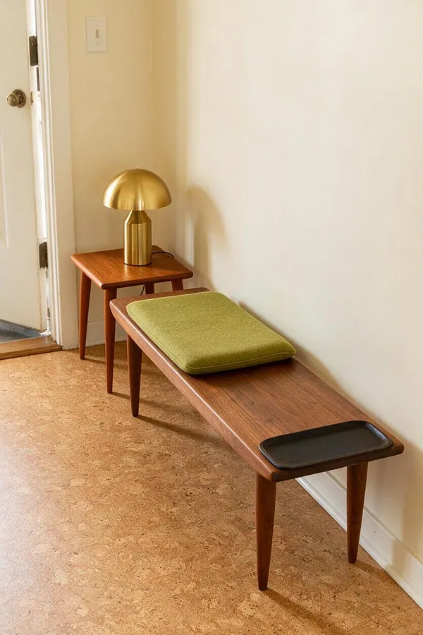 A narrow entryway with a long walnut bench, tapered legs, and a green upholstered cushion, paired with a small matching side table holding a brass mushroom lamp. Cream walls and cork flooring create a warm, minimalist mid-century modern look.