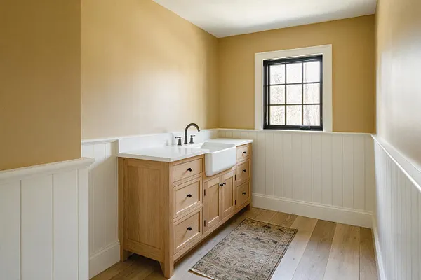 A farmhouse-style powder room with warm tan walls, white beadboard wainscoting, and a light oak vanity topped with a white countertop. A black-framed window brings in daylight above the deep apron-front sink, and a muted vintage-style rug softens the light wood floor.