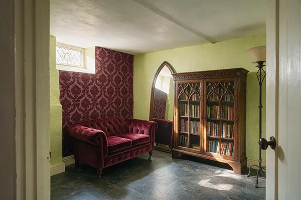 A small Gothic-style basement room with a burgundy velvet loveseat, a carved wooden bookcase filled with books, and an arched standing mirror. The walls mix pale green paint with deep burgundy damask wallpaper, while a slate or stone-look floor and a tall black floor lamp complete the moody, old-world atmosphere.