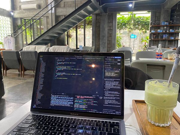 A laptop displaying code sits on a table in a quiet cafe, accompanied by a green beverage.