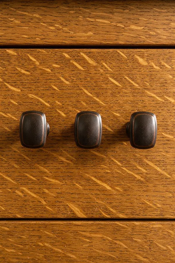 A close-up of a Craftsman-style wooden cabinet front in warm golden oak, showing distinctive linear grain and three dark bronze knob pulls arranged horizontally across the center. The hardware and wood texture create a simple, handcrafted look.