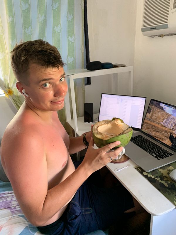 A photo of a shirtless man with glasses relaxing on a gray couch in a room filled with books and a fan.