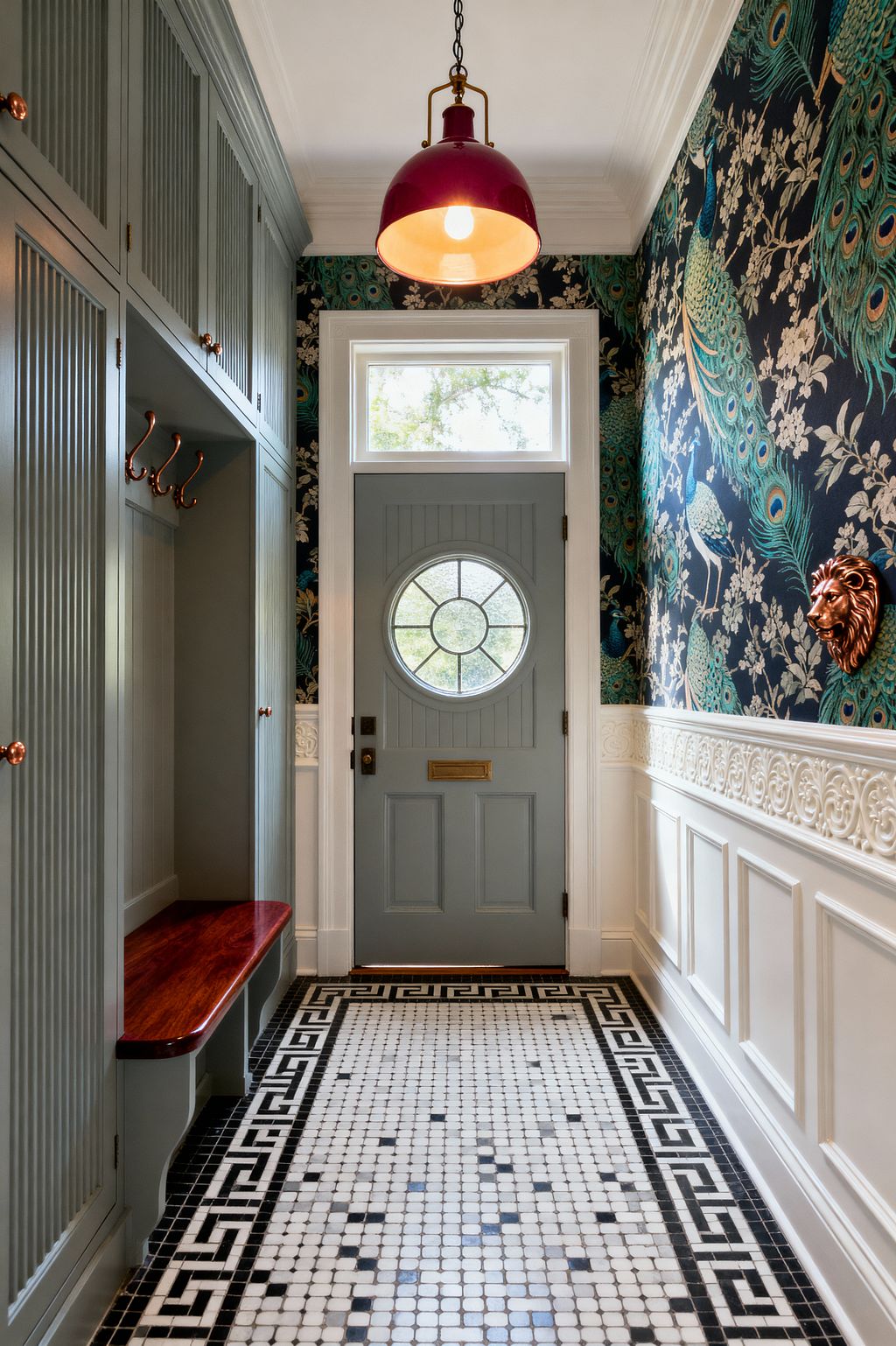 A narrow Victorian mudroom with a blue-gray paneled door featuring a circular leaded-glass window and a transom above. Black-and-white mosaic tile with a Greek key border leads past sage-gray built-in lockers with copper hooks and a glossy wood bench. Navy wallpaper with teal-and-gold peacocks sits over ornate white wainscoting, while a copper lion-head hook and a berry-red pendant light add warm metallic accents.