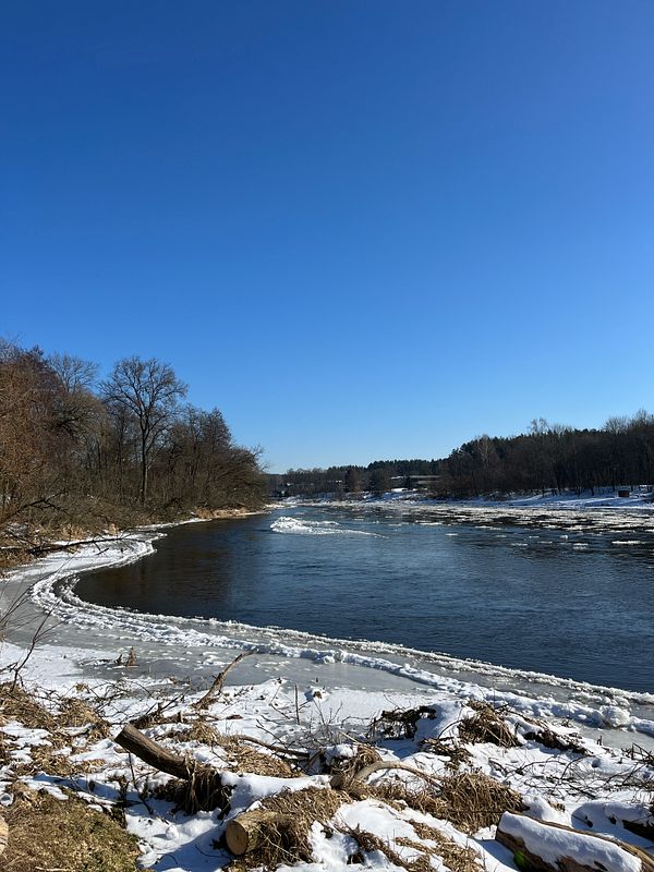 A serene winter landscape featuring a river bordered by snow and trees under a clear blue sky.