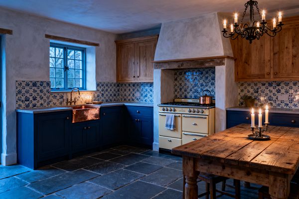 A cozy farmhouse kitchen shows deep navy Shaker cabinets with a hammered copper apron-front sink and brass bridge faucet beneath a blue-trimmed window. Blue-and-white floral ceramic tiles form the backsplash around a cream enamel range set in a plaster hood with a wood beam; honey-toned oak cupboards, a chunky reclaimed wood table, a wrought-iron candle chandelier, and dark slate flagstone floors complete the rustic, candlelit scene.