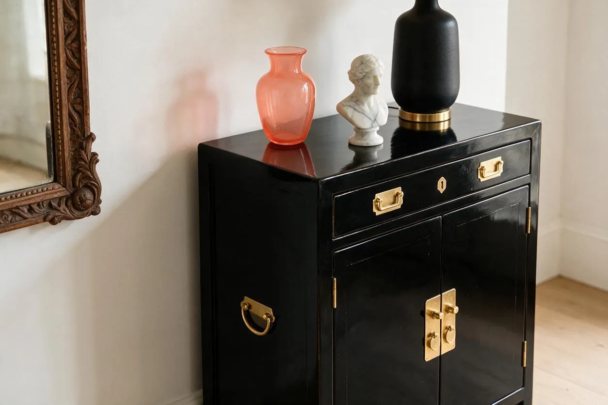 A glossy black cabinet with brass hardware stands against a light wall, styled with a coral glass vase, a small white bust, and a matte black vase on top. An ornate carved mirror frame is partially visible on the left, adding to the eclectic mix of finishes and objects.