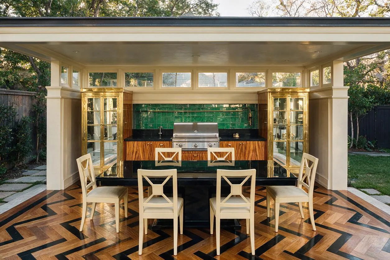 An outdoor backyard kitchen pavilion with a glossy black counter, brass-trimmed glass cabinets, a stainless steel grill, and emerald green tile backsplash. Four cream dining chairs sit on a bold herringbone and chevron wood floor, under a flat-roofed structure with cream columns and clerestory windows.