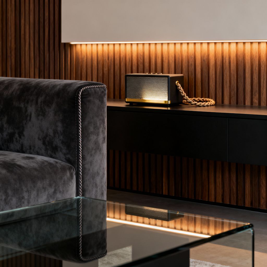 Close-up of a contemporary media room featuring vertical walnut slat wall paneling with warm LED backlighting, a floating matte-black media console, and a compact speaker with brass trim and a braided rope handle. In the foreground, a charcoal velvet sofa with piped edges sits beside a clear glass coffee table over polished concrete flooring, creating a moody, refined atmosphere.