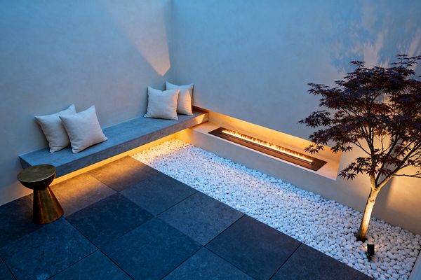 A compact walled patio at dusk features a floating concrete bench with light linen pillows along a smooth plaster corner. Warm LED strips glow beneath the seat and within a narrow recessed linear fire set into the low wall. Large charcoal stone pavers edge a bed of crisp white river pebbles, where a small Japanese maple is up-lit from below. A sculptural brass side table sits beside the bench, emphasizing the minimalist palette and clean geometry.