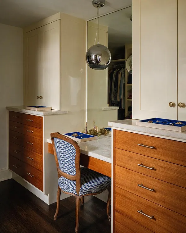 A stylish eclectic closet featuring built-in cream cabinetry with wood veneer drawers, a marble vanity counter, and a large mirrored wall. A chrome pendant light hangs above a small dressing table with a patterned upholstered chair, and blue jewelry trays add a colorful accent.