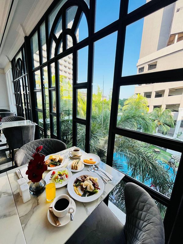 A beautifully arranged breakfast spread is displayed on a marble table beside large windows overlooking a pool and palm trees.