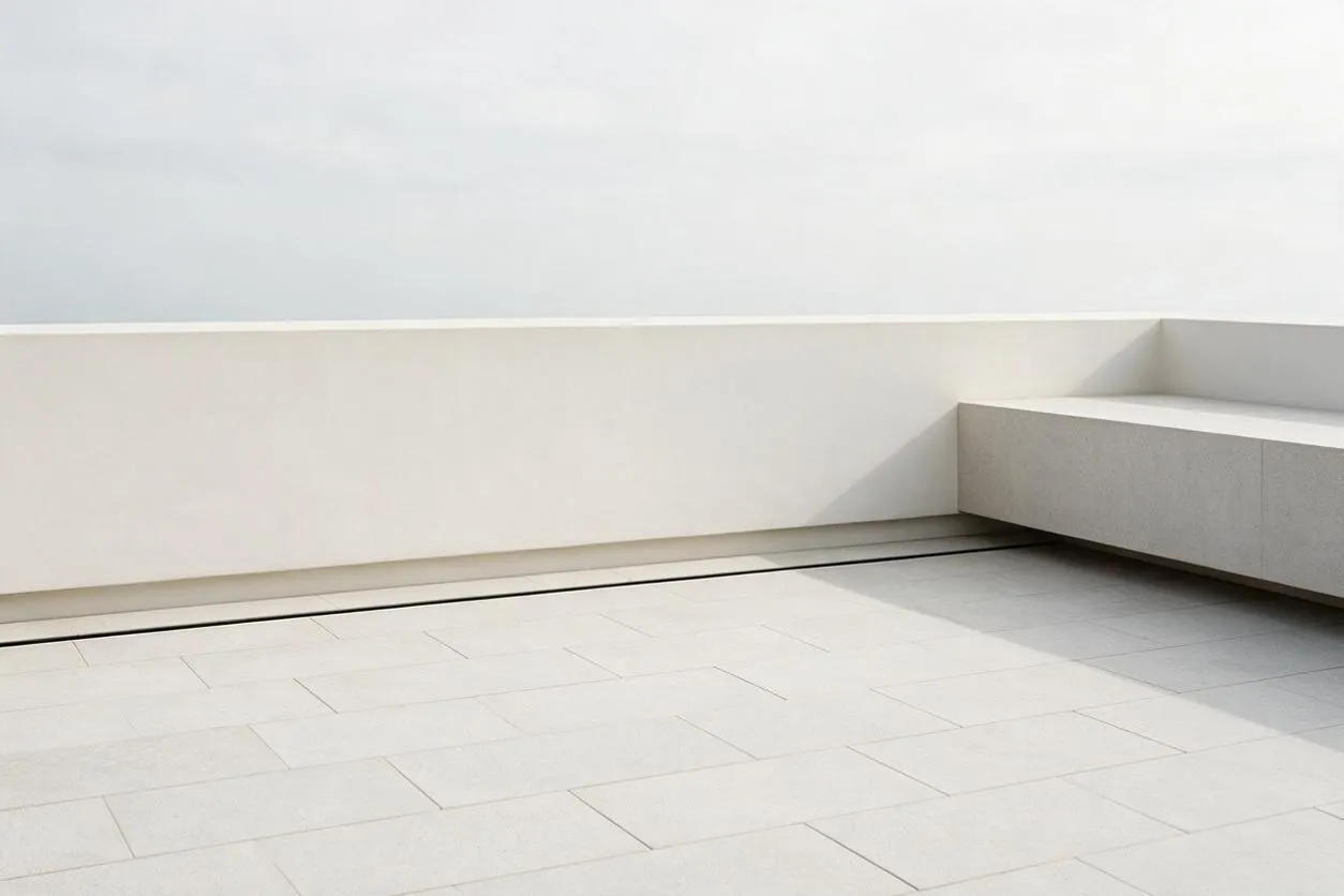 A sparse minimalist rooftop terrace with large pale tiled flooring, a low white perimeter wall, and a built-in bench or ledge in light gray on the right. The space is open to a bright sky, with soft shadows emphasizing the simple geometry and uncluttered design.