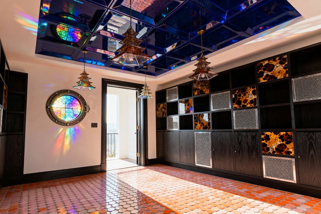 A maximalist mudroom features a cobalt mirrored ceiling in a grid, reflecting light from three tiered amber-glass pendants. Along one wall, dark-stained built-ins mix open cubbies, tortoiseshell-patterned resin panels, and perforated metal doors above matte wood cabinets. A round porthole of multicolor stained glass casts rainbow reflections across warm white walls and a gradient floor of orange and blush penny tiles, while a black-trimmed doorway opens to a balcony.