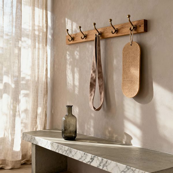 A sunlit corner of a home gym shows a warm taupe plaster wall with an oak peg rail and brass double hooks holding a neutral fabric strap and a perforated wooden paddle. Below, a cantilevered concrete bench with a white marble edge supports a smoky glass bottle while sheer ecru linen curtains filter daylight, casting soft shadows that emphasize the room’s clean lines and tactile finishes.