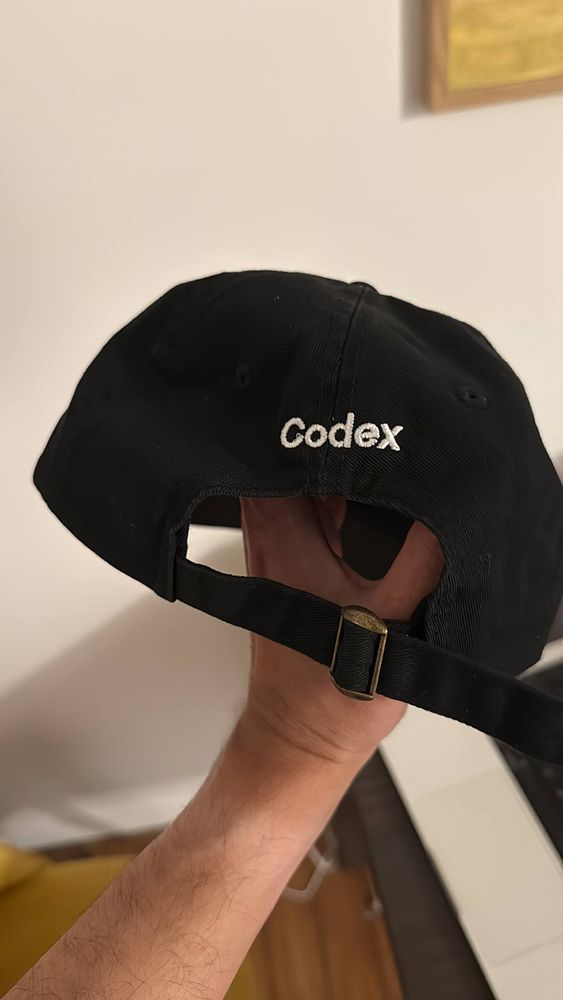 A black baseball cap with an embroidered white 'OpenAI' logo on the front is shown on a light wooden surface.