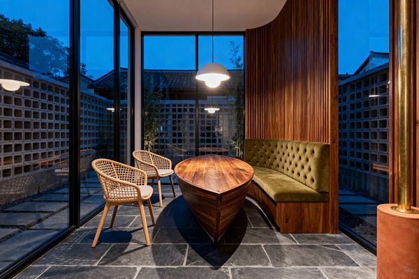 A mid-century modern dining nook with a sculptural, boat-shaped walnut table sits centered between a curved, tufted olive velvet banquette set against vertical walnut slat paneling and two woven rattan armchairs. Large floor-to-ceiling windows with black frames reveal breeze-block garden walls at dusk, while a white globe pendant casts warm light onto charcoal slate flooring, brass detailing, and the rich wood surfaces, creating a cozy contrast to the cool blue exterior.
