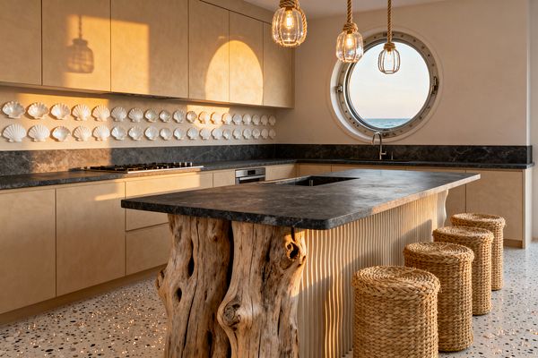 A warm coastal kitchen features sandy matte cabinets, charcoal stone countertops, and a large round porthole window overlooking the sea. A sculptural island with a fluted base and driftwood supports pairs with woven rattan stools, rope-wrapped glass pendants, a terrazzo floor, a gas cooktop, and a horizontal band of white ceramic scallop shells across the backsplash, all washed in golden sunset light.