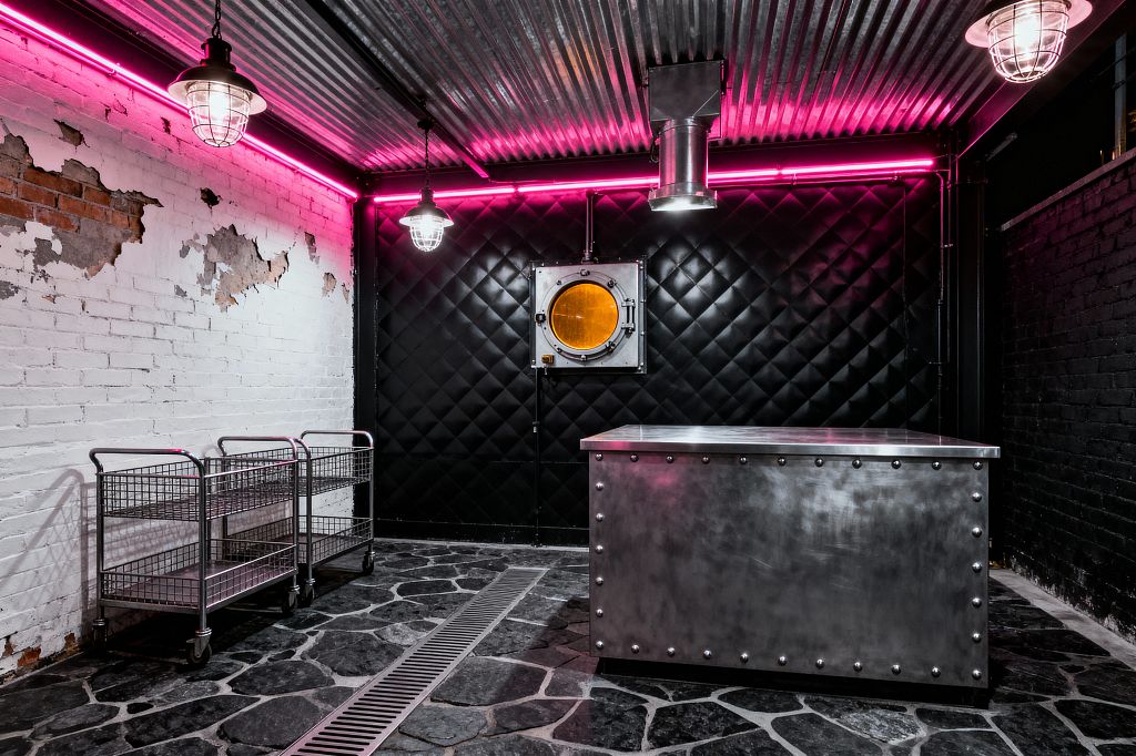 An industrial laundry room with a corrugated metal ceiling and cage-shaded pendants under a perimeter of magenta neon. A glossy black, diamond-quilted wall with a round amber porthole contrasts distressed white brick; a riveted stainless-steel island sits on dark flagstone with a linear floor drain, while two chrome wire rolling carts line the left wall.
