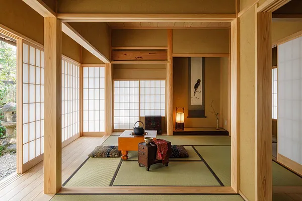 A tranquil Japanese Zen-style sunroom with light wood posts and beams, shoji screen windows, and tatami mats covering the floor. In the center sits a low tea table with a teapot and cushions, while a softly lit alcove with a hanging scroll and lantern adds a calm focal point.