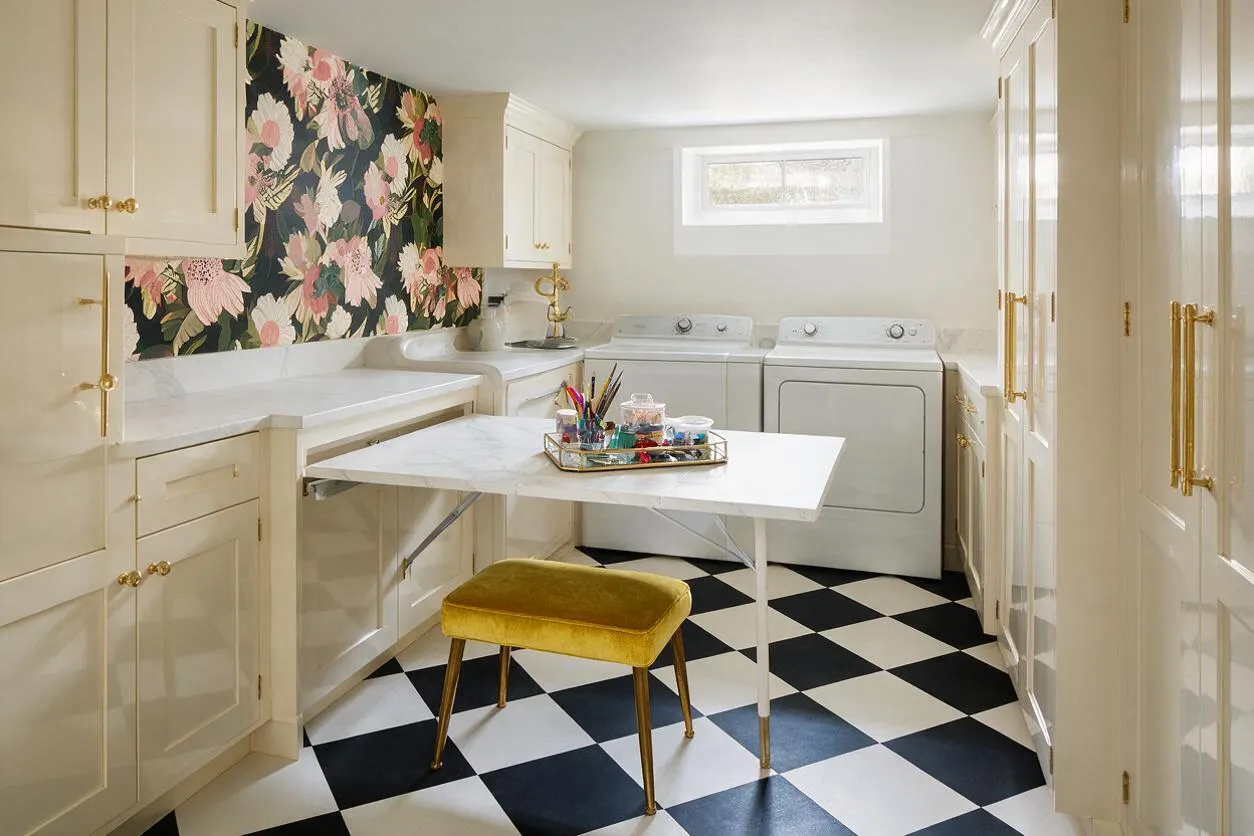 A compact basement laundry room with cream cabinetry, brass hardware, a black-and-white checkered floor, and a white folding table centered in the space. A bold floral wallpaper accent wall adds Hollywood Regency flair, while a mustard velvet stool, white appliances, and a small high window brighten the room.
