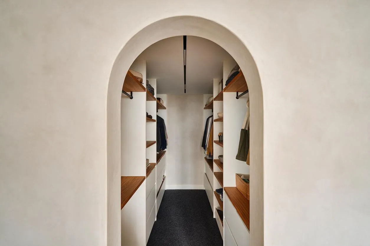 A narrow modern walk-in closet is framed by a soft arched doorway and lined with white built-in cabinetry. Warm wood shelves and hanging rods run along both sides, with a dark floor runner and a minimalist ceiling light creating a clean, streamlined look.