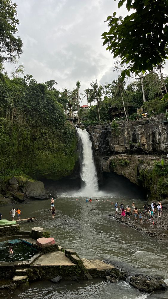 A scenic waterfall in Bali surrounded by lush greenery and visitors enjoying the water.