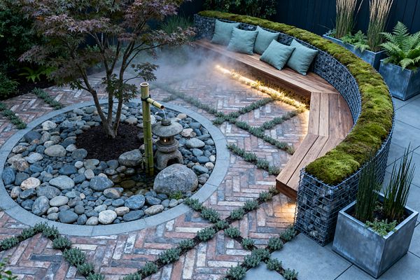 An intimate patio centers on a circular river-stone bed with a Japanese maple, a bamboo water spout, and a small stone lantern beside a shallow pool. Surrounding it are reclaimed herringbone brick pavers divided by planted joints. A curved gabion bench with a wooden seat and moss-topped back includes a warm linear LED light. Concrete planters with ferns and upright reeds sit against a dark fence while a soft mist drifts through the scene.