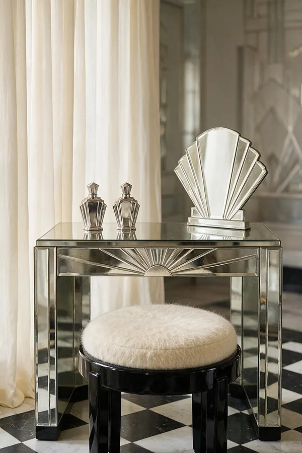 An Art Deco-inspired vanity vignette with a mirrored glass table featuring sunburst detailing, two faceted perfume bottles, and a fan-shaped mirror accessory on top. A round black stool with a cream furry cushion sits in front, set against sheer ivory curtains and a black-and-white checkered floor.