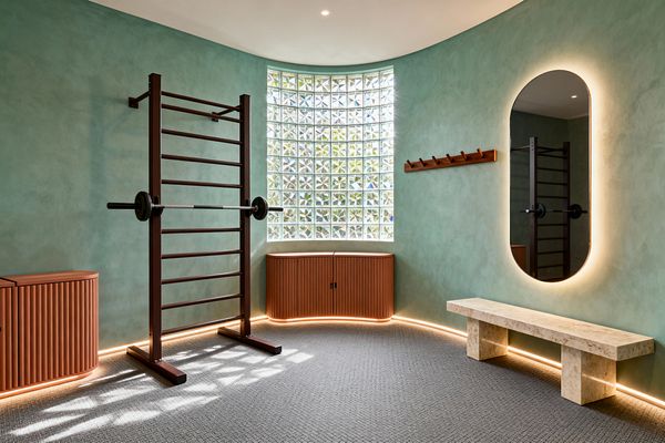 A contemporary home gym with sage-green limewash walls features a dark stall bar holding a barbell, a tall oval mirror with halo backlighting, and a travertine bench. A curved glass-block window brings in diffused daylight above fluted terracotta storage, echoed by a matching side unit and a wooden peg rail. Perimeter LED strips wash the base of the walls, and a gray textured floor and white ceiling with recessed spots complete the calm space.